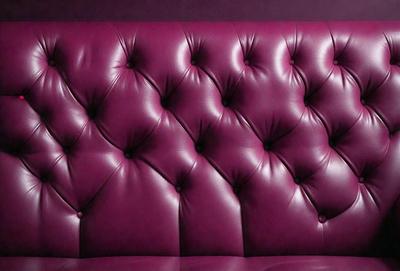 The primary subject of the image is a purple couch with a leather-like texture. The couch is situated in a room with a purple wall, which adds to the overall atmosphere of the scene. The visual style of the image is a close-up of the couch, showcasing its design and texture. The colors and mood of the image are predominantly purple, creating a cozy and inviting ambiance.
