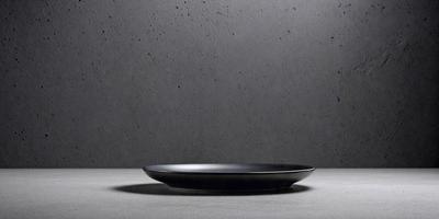 The primary subject of the image is a black plate placed on a table. The setting is a grey cement wall, which serves as the backdrop for the plate. The visual style is a photo, capturing the plate's placement on the table. The colors in the image are predominantly black and grey, creating a mood of simplicity and minimalism.