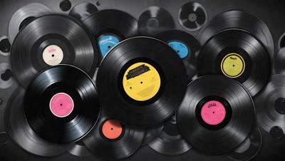 The primary subject of the image is a collection of various colored vinyl records. The records are arranged in a visually appealing manner, creating a sense of harmony and unity. The setting is a black background, which provides a contrasting and dramatic backdrop for the records. The visual style of the image is a combination of photo and digital art, as the records are displayed in a way that resembles a painting or a collage. The colors and mood of the image are vibrant and lively, with the different colors of the records creating a sense of excitement and energy.