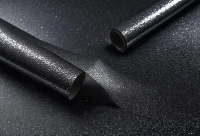 The primary subject of the image is a roll of glittery material, which is placed on a black surface. The setting appears to be a dark room, and the visual style is a close-up of the glittery material. The colors and mood of the image are predominantly dark and mysterious, with the glittery material adding a touch of sparkle and interest to the scene.
