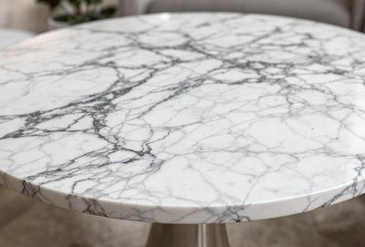 The primary subject of the image is a white marble table. The table is placed on a white surface, which could be a table or a countertop. The visual style of the image is a photo, showcasing the marble table and its texture. The colors and mood of the image are predominantly white, giving it a clean and elegant appearance.