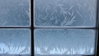 The primary subject of the image is a window with frost and ice on it. The setting is a cold, winter environment. The visual style is a photo of the window with frost and ice on it. The colors and mood are predominantly cold and icy, with the frost and ice creating a striking visual effect.