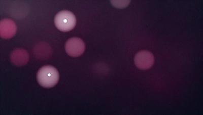 The primary subject of the image is a purple background with a pattern of white dots. The setting appears to be a dark room, and the visual style is a close-up of the purple background with the white dots. The colors and mood of the image are predominantly purple and dark, creating a moody and atmospheric scene.