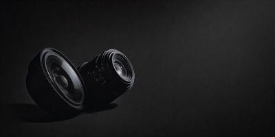 The primary subject of the image is a close-up of a camera lens. The lens is placed on a black surface, which serves as the setting or background. The visual style of the image is a black and white photo, giving it a classic and timeless appearance. The colors and mood of the image are predominantly dark, with the lens being the focal point and the black background providing a sense of depth and contrast.