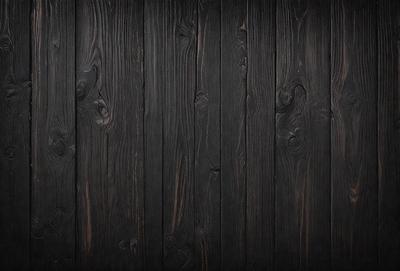 The primary subject of the image is a wooden wall with a black color. The setting is a room with a black background. The visual style is a close-up of the wooden wall, which gives a sense of texture and depth. The colors and mood of the image are dark and moody, creating a sense of mystery or drama.