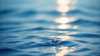 The primary subject of the image is a small water drop, which is the main focus of the scene. The setting is a body of water, possibly an ocean, with a blue background. The visual style of the image is a photo, capturing the water drop in its natural environment. The colors in the image are predominantly blue, creating a calm and serene atmosphere.