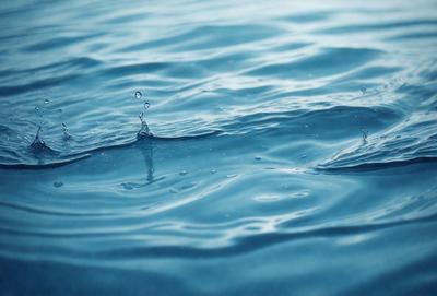 The primary subject of the image is a body of water, which appears to be a large lake or ocean. The water is blue and has a few droplets of water floating on its surface. The setting is a vast body of water, with no visible land or other objects in the background. The visual style of the image is a realistic representation of the water, with no indication of it being a photo, digital art, illustration, drawing, pattern, texture, or clipart. The colors and mood of the image are serene and peaceful, with the blue water and the floating droplets creating a calming atmosphere.