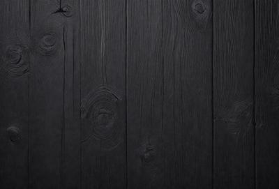 The primary subject of the image is a wooden wall with a black surface. The setting is a room with a wooden floor. The visual style is a close-up of the wooden wall, showcasing its texture and details. The colors and mood of the image are dark and moody, with the black wooden wall and floor creating a dramatic atmosphere.