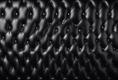 The primary subject of the image is a black leather couch. The setting is a black background, which creates a striking contrast with the couch. The visual style is a close-up of the couch, showcasing its texture and details. The colors and mood of the image are predominantly black, giving it a sleek and modern appearance.