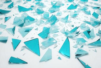 The primary subject of the image is a collection of broken glass pieces, predominantly blue in color. The setting is a white surface, possibly a table or a wall, where the broken glass pieces are scattered. The visual style of the image is a close-up of the broken glass pieces, showcasing their intricate patterns and textures. The colors and mood of the image are predominantly blue, with a sense of fragility and impermanence due to the broken nature of the glass pieces.