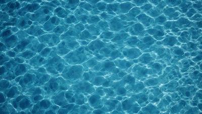 The primary subject of the image is a large body of water, which appears to be a swimming pool. The water is blue and has a rippling effect, creating a visually appealing and serene atmosphere. The setting is a swimming pool, and the visual style is a photo. The colors and mood of the image are predominantly blue, creating a sense of calmness and relaxation.