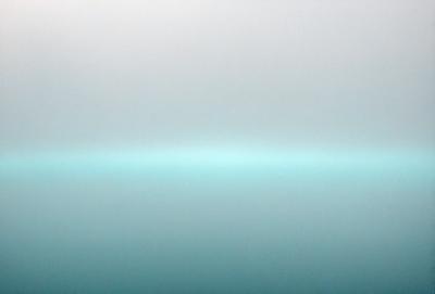 The primary subject of the image is a large body of water, which appears to be a lake. The setting is a foggy, cloudy day, creating a somewhat mysterious atmosphere. The visual style of the image is a photo, capturing the beauty of the lake on a foggy day. The colors and mood of the image are predominantly blue, with the water and the sky creating a serene and peaceful ambiance.