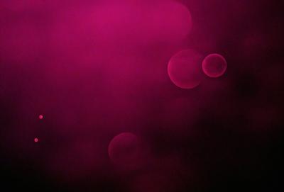 The primary subject of the image is a pink background with three red circles floating in the air. The setting appears to be a dark, foggy environment, creating a mysterious and atmospheric mood. The visual style of the image is a combination of photo and digital art, as it captures the red circles in a somewhat surreal manner. The colors and mood of the image are predominantly pink and red, with a hint of darkness and intrigue.