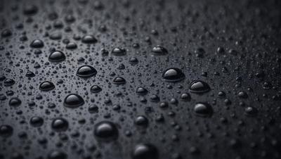 The primary subject of the image is a close-up of a black surface with numerous small droplets of water on it. The setting is a dark background, and the visual style appears to be a photo. The colors and mood of the image are predominantly dark, with the droplets of water being the main focus.