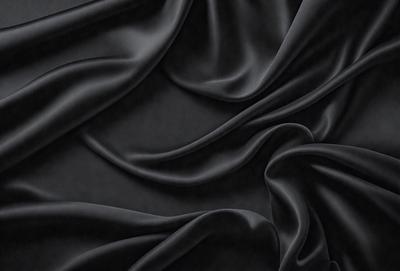The primary subject of the image is a close-up of a black fabric, possibly a piece of clothing or a curtain. The setting is a dark background, which emphasizes the fabric's texture and details. The visual style appears to be a photo, as it captures the fabric's intricate patterns and textures. The colors and mood of the image are predominantly dark, with the black fabric serving as the focal point.