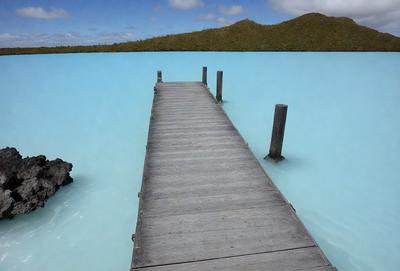 The primary subject of the image is a wooden pier extending out into a large body of water. The pier is surrounded by a serene environment, with a beautiful blue lake and lush greenery in the background. The visual style of the image is a photo, capturing the natural beauty of the scene. The colors and mood of the image are predominantly blue and calm, creating a peaceful atmosphere.