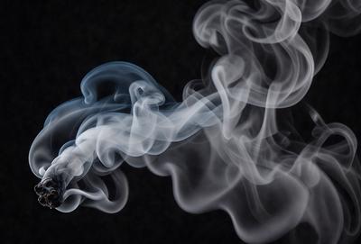 The primary subject of the image is a cigarette, which is the main focus of the scene. The setting is a dark background, which creates a moody atmosphere. The visual style is a close-up of the cigarette, emphasizing its details. The colors in the image are predominantly shades of gray, which contribute to the overall mood of the scene.