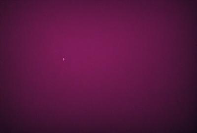 The primary subject of the image is a single moon, which is located in the middle of the purple sky. The setting is a dark, cloudy sky, and the visual style appears to be a photo. The dominant colors in the image are purple and black, creating a moody and atmospheric scene.