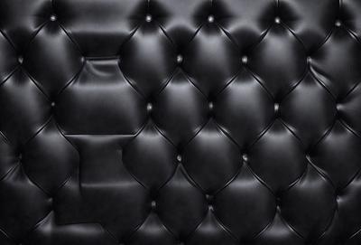 The primary subject of the image is a black leather couch. The setting is a black background, which emphasizes the couch's color and texture. The visual style is a close-up of the couch, showcasing its details. The colors and mood of the image are dark and moody, with the black leather couch being the main focal point.