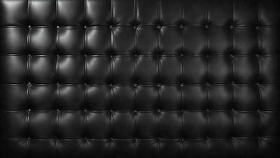 The primary subject of the image is a black leather couch. The setting is a black background, which creates a striking contrast with the couch. The visual style is a close-up of the couch, showcasing its texture and details. The colors and mood of the image are predominantly black, giving it a sleek and modern appearance.