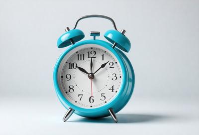 The primary subject of the image is a blue alarm clock with a white face. The clock is placed on a white surface, which serves as the background. The visual style of the image is a photo, capturing the details of the alarm clock. The colors and mood of the image are predominantly blue and white, creating a clean and minimalist appearance.