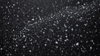 The primary subject of the image is a snowy scene with a black background. The setting appears to be a nighttime environment, possibly in a city. The visual style of the image is a photo, capturing the snowflakes in motion. The dominant colors in the image are white and black, creating a serene and wintry atmosphere.