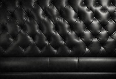 The primary subject of the image is a black leather couch with a black and white pattern. The couch is positioned against a black wall, which serves as the setting or background. The visual style of the image is a black and white photo, giving it a classic and timeless appearance. The dominant colors in the image are black and white, creating a moody and sophisticated atmosphere.