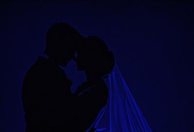 The primary subject of the image is a bride and groom, dressed in their wedding attire, standing close to each other. They are embracing each other, with the bride wearing a veil. The setting is a dark room, possibly a nighttime wedding reception. The visual style is a black and white photo, which adds a classic and timeless feel to the image. The dominant colors are black and white, and the overall atmosphere is intimate and romantic.