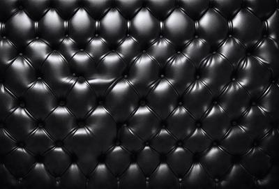 The primary subject of the image is a black leather couch. The setting is a black background, which creates a striking contrast with the couch. The visual style is a close-up of the couch, showcasing its texture and details. The colors and mood of the image are predominantly black, giving it a sleek and modern appearance.