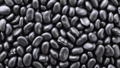 The primary subject of the image is a large pile of black beans. The beans are arranged in a visually appealing manner, creating a sense of depth and texture. The setting is a black background, which emphasizes the beans and their arrangement. The visual style is a close-up of the beans, showcasing their details and texture. The colors and mood of the image are predominantly black, giving it a bold and striking appearance.