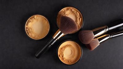 The primary subject of the image is a collection of four makeup brushes, each with different colors and sizes. They are placed on a black surface, which serves as the setting or background. The visual style of the image is a close-up of the brushes, showcasing their details and colors. The colors and mood of the image are predominantly dark, with the black surface and the makeup brushes creating a moody atmosphere.