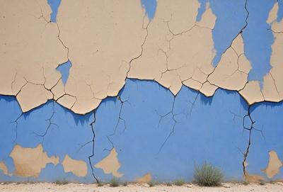The primary subject of the image is a blue wall with a cracked and chipped appearance. The setting is a desert-like environment, with a dirt surface visible in the background. The visual style is a photo, capturing the natural texture and colors of the wall. The dominant colors are blue and brown, with the blue wall contrasting against the brown dirt. The overall atmosphere is one of wear and tear, possibly due to the harsh desert conditions.
