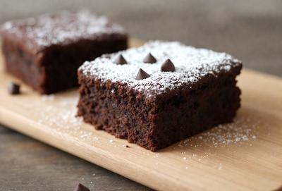 The image features a delicious chocolate cake with white frosting and chocolate chips on top. The cake is placed on a wooden cutting board, which is resting on a dining table. The cake is the primary subject, and the setting is a cozy home environment. The visual style is a close-up of the cake, showcasing its mouth-watering appearance. The colors and mood of the image are warm and inviting, with the rich chocolate and white frosting creating a sense of indulgence.