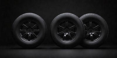 The primary subject of the image is a set of four black tires. They are placed on a black background, which creates a striking contrast with the tires. The visual style of the image is a photo, capturing the details of the tires. The colors and mood of the image are dark and dramatic, emphasizing the tires' shape and design.