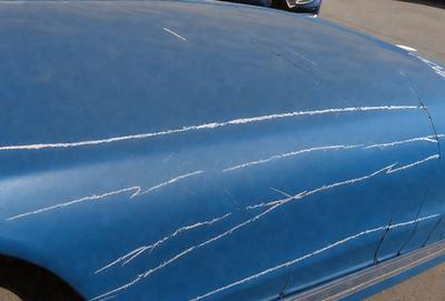 The primary subject of the image is a blue car with a scratched surface. The setting is a parking lot, and the visual style appears to be a photo. The car has a blue leather-like material, and the dominant colors are blue and white. The overall atmosphere of the image is somewhat damaged or worn, possibly due to the scratches on the car's surface.