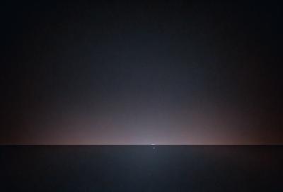 The primary subject of the image is a lone light in the middle of a dark, starry sky. The setting is a vast, open space with no visible land or other objects. The visual style is a black and white photo, which adds a sense of timelessness and mystery to the scene. The colors and mood are predominantly dark and moody, with the lone light serving as a focal point in the otherwise empty and expansive sky.
