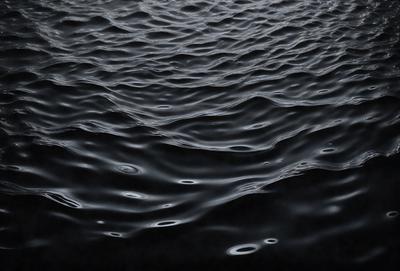 The primary subject of the image is a large body of water, possibly an ocean or a lake. The water appears to be dark and has a few ripples on its surface. The setting is a vast expanse of water, with no visible land or other objects in the background. The visual style of the image is a black and white photo, which adds a sense of depth and contrast to the scene. The overall atmosphere of the image is calm and serene, with the water's surface providing a sense of tranquility.