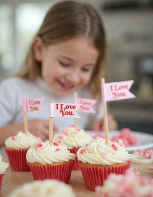 The primary subject of the image is a young girl smiling and holding a pink flag. She is surrounded by cupcakes, with three of them placed in front of her and another one on the right side of the image. The setting appears to be a kitchen or a similar indoor space. The visual style of the image is a photo, capturing the girl's joyful expression and the festive atmosphere created by the cupcakes. The colors and mood of the image are predominantly pink, which adds a