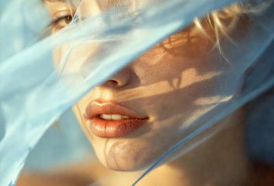 The primary subject of the image is a woman with a beautiful smile. She is wearing lipstick and has a blue veil covering her face. The setting is outdoors, with a blue sky visible in the background. The visual style is a close-up of the woman's face, emphasizing her features and expression. The colors in the image are predominantly blue, creating a calm and serene atmosphere.