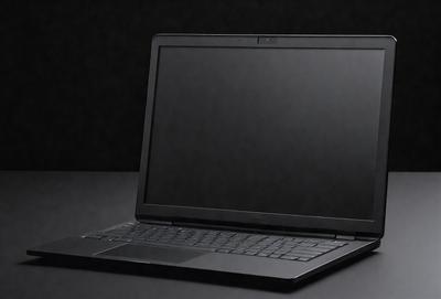 The primary subject of the image is a laptop computer. The laptop is sitting on a table, and it is turned on. The setting is a dark room, which creates a moody atmosphere. The visual style is a photo, capturing the laptop in its current state. The colors in the image are predominantly black, with the laptop screen being the main source of light and color.