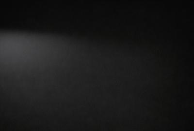 The primary subject of the image is a bright light source, which appears to be a light bulb or a spotlight. The light source is located in the upper left corner of the image. The setting is a dark room, with the light source being the only source of illumination. The visual style of the image is a black and white photo, which adds a dramatic and artistic touch to the scene. The colors and mood of the image are predominantly dark, with the bright light source standing out as the focal