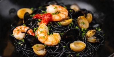 The primary subject of the image is a plate of food, featuring a combination of shrimp, scallops, and pasta. The dish is presented on a black surface, which contrasts with the vibrant colors of the seafood and pasta. The visual style of the image is a close-up, showcasing the details of the food. The colors and mood of the image are lively and appetizing, with the shrimp and scallops adding a touch of freshness to the