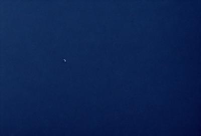 The primary subject of the image is the moon, which is seen in the sky. The setting is a vast, dark blue sky, with no clouds or other objects visible. The visual style is a photo, capturing the moon in its natural environment. The colors and mood of the image are predominantly dark and moody, with the moon being the only light source in the scene.