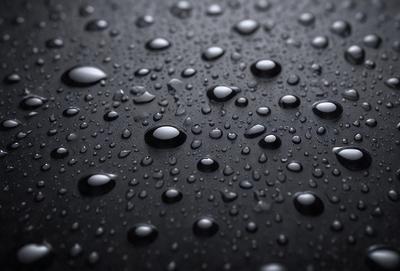 The primary subject of the image is a close-up of a black surface with numerous droplets of water on it. The setting is a dark background, and the visual style appears to be a photo. The colors and mood of the image are predominantly dark, with the droplets of water being the main focus.