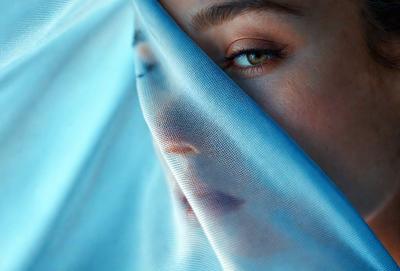 The primary subject of the image is a woman with a blue veil covering her face. She is looking at the camera with a mysterious expression. The setting is a room with a blue background, and the visual style appears to be a photo. The woman's face is the main focus of the image, and the overall atmosphere is somewhat mysterious and intriguing.