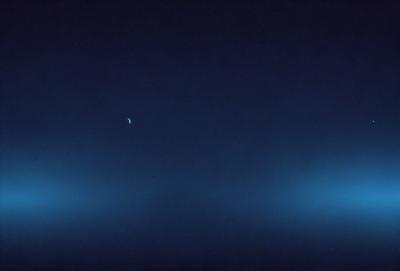 The primary subject of the image is a moon, which is visible in the sky. The setting is a dark, cloudy night with a blue sky. The visual style is a photo, capturing the moon and the night sky. The colors and mood of the image are predominantly dark and moody, with the blue sky providing a contrasting element.