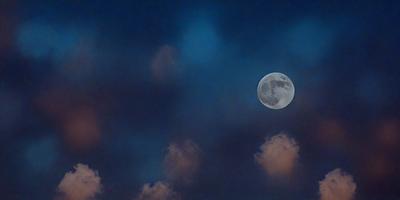 The primary subject of the image is a full moon, which is prominently displayed in the sky. The setting is a cloudy night sky, with the moon being the main focus. The visual style of the image is a photo, capturing the beauty of the moon against the backdrop of the cloudy sky. The colors and mood of the image are predominantly dark and moody, with the moon being the only source of light in the scene.
