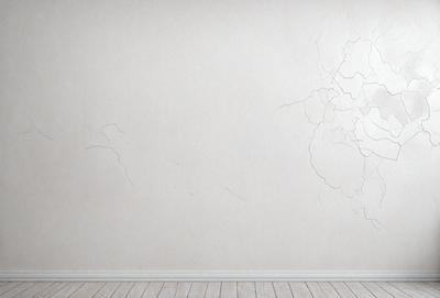 The primary subject of the image is a white wall with a cracked and chipped appearance. The setting is a room with a wooden floor. The visual style is a photo, and the dominant colors are white and grey, creating a somewhat dull and aged atmosphere.