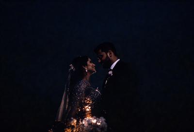The primary subject of the image is a bride and groom, dressed in their wedding attire, standing close to each other and kissing. The setting is a nighttime outdoor wedding ceremony, with a dark sky in the background. The visual style is a combination of a photo and digital art, creating a unique and artistic representation of the couple's special moment. The colors and mood of the image are warm and romantic, reflecting the joy and love shared by the newlyweds.