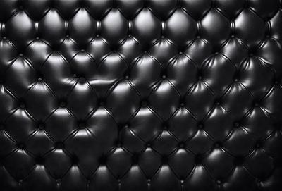 The primary subject of the image is a black leather couch. The setting is a black background, which creates a striking contrast with the couch. The visual style is a close-up of the couch, showcasing its texture and details. The colors and mood of the image are predominantly black, giving it a sleek and modern appearance.